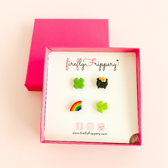 Set of four stud earrings - a four leaf clover, pot of gold, half a rainbow and three leafed shamrock - on a fireflyFrippery branded jewelry card in a hot pink square box sitting on the gift box lid