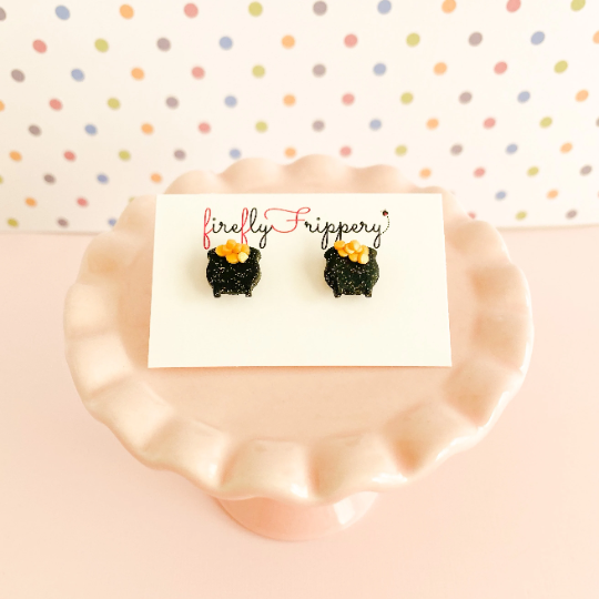 Pair of pot of gold stud earrings on a fireflyFrippery branded jewelry card resting on a miniature, soft pink cupcake stand in front of a pastel polka-dot background