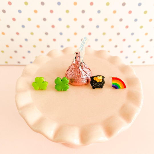 Set of four stud earrings - a four leaf clover, pot of gold, half a rainbow and three leafed shamrock - arranged in a line beside and in front of a Hershey's kiss, resting on a soft pink miniature cupcake stand in front of a pastel polka-dot background