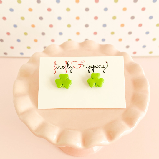 Pair of bright green three leafed shamrock stud earrings on a fireflyFrippery branded jewelry card resting on a miniature, soft pink cupcake stand in front of a pastel polka-dot background
