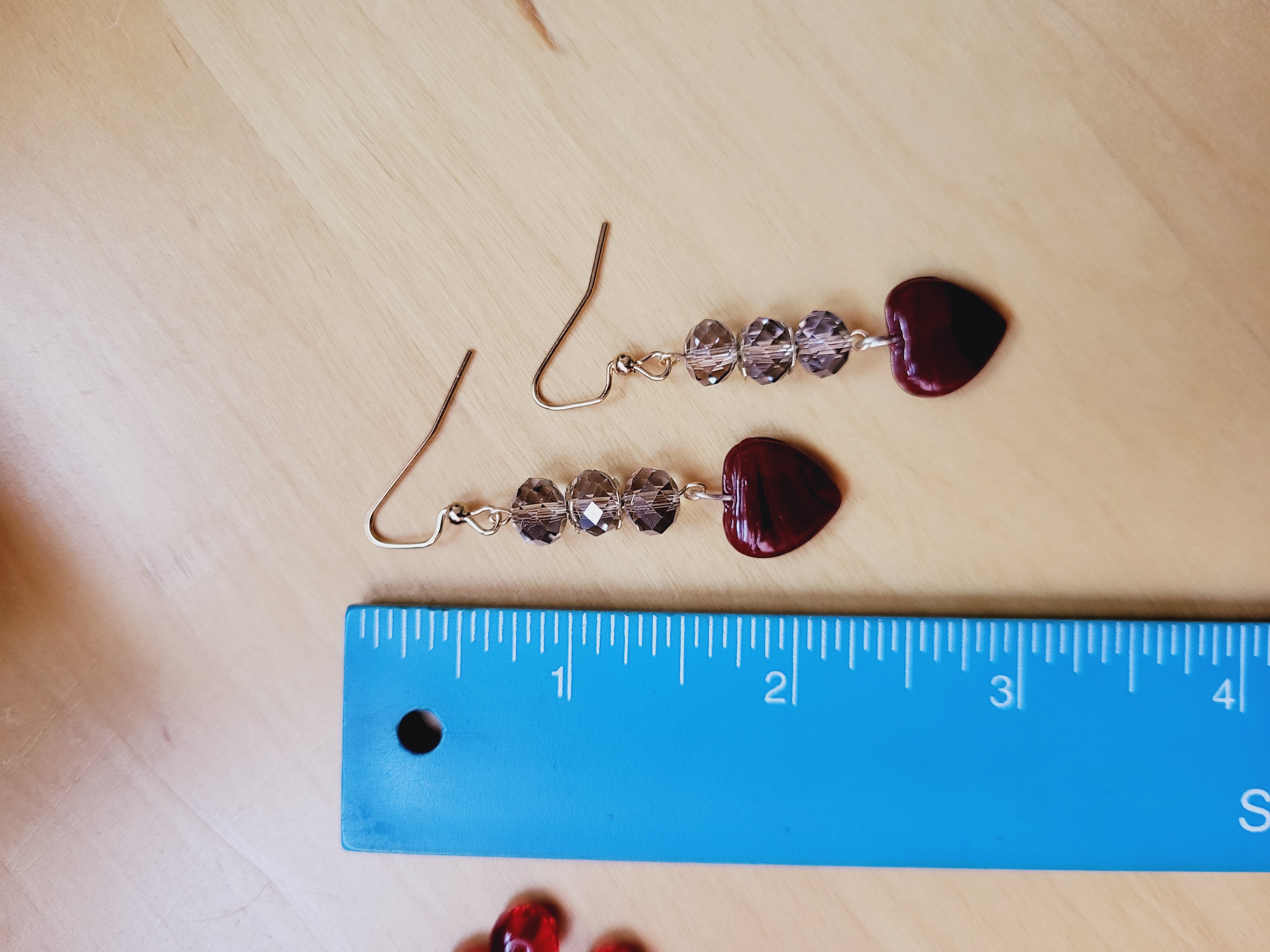 Light Gray Beaded Dangle Earrings with Medium Red Banded Heart Charm lay on a table beside a ruler.  They measure approx. 2.25 inches long.