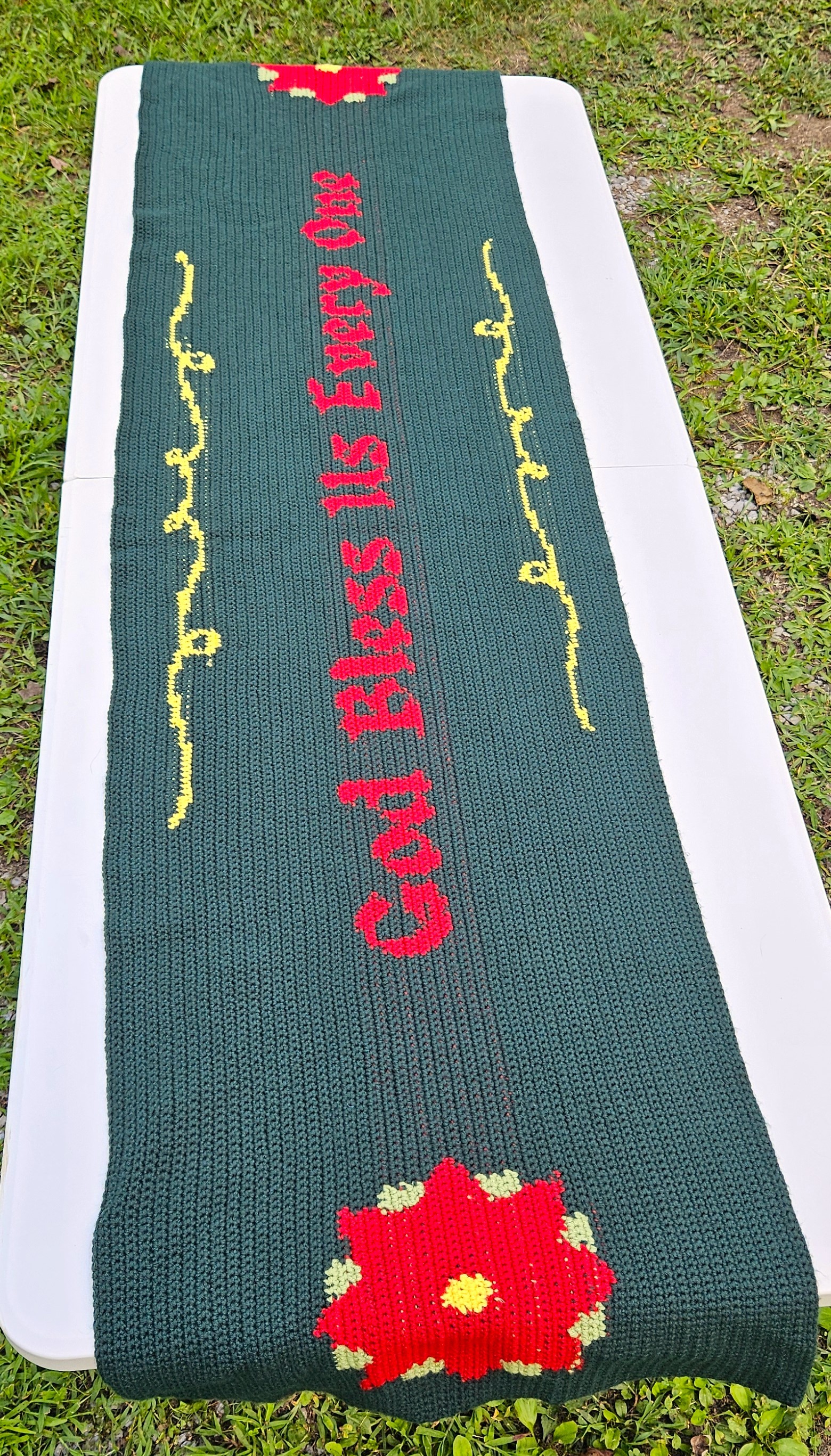 A dark green and red table runner with God Bless Us Every One in the center, poinsettias on either end. And yellow swirls around the quote. A dark green and red table runner with God Bless Us Every One in the center, poinsettias on either end. And yellow swirls around the quote.