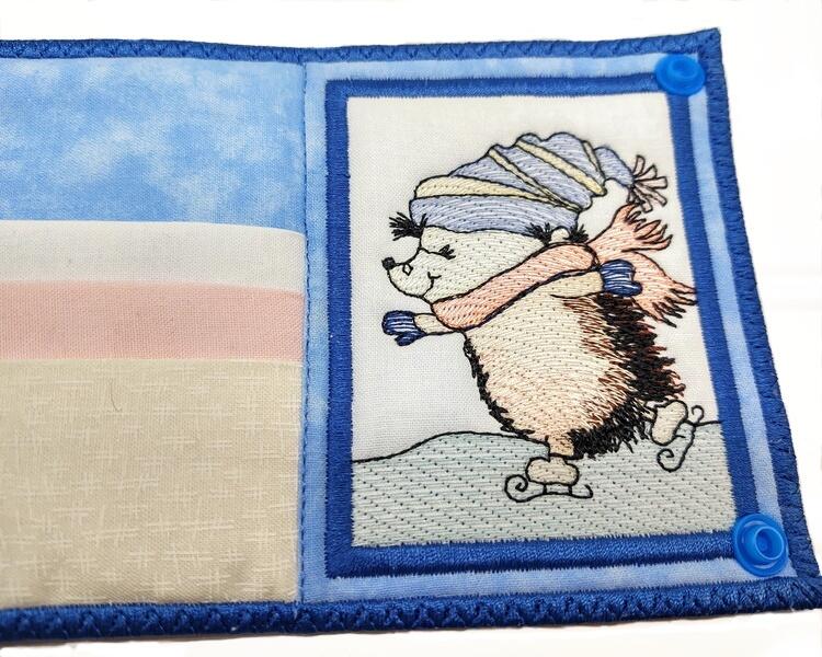 A medium blue interior and exterior, with a white, pink, and beige tea bag pockets. The hedgehog on the right side is done in machine embroidery.  He's brown with dark brown accents. He has a light blue and beige hat and a pink scarf.