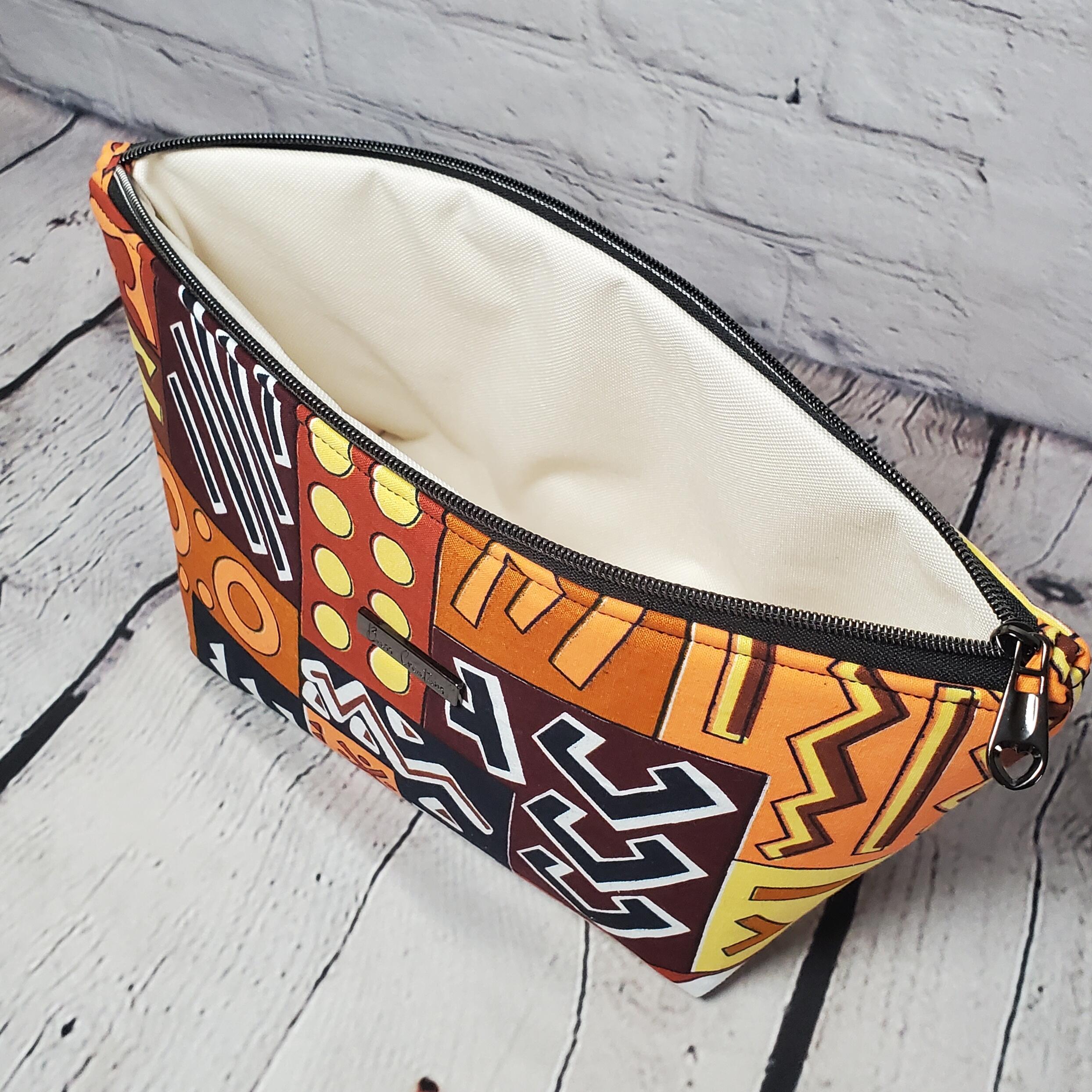 Top view of a brown orange and yellow geometric African print cosmetic bag with a gunmetal zipper, ivory interior waterproof canvas lining and a gunmetal Bass Creations logo.