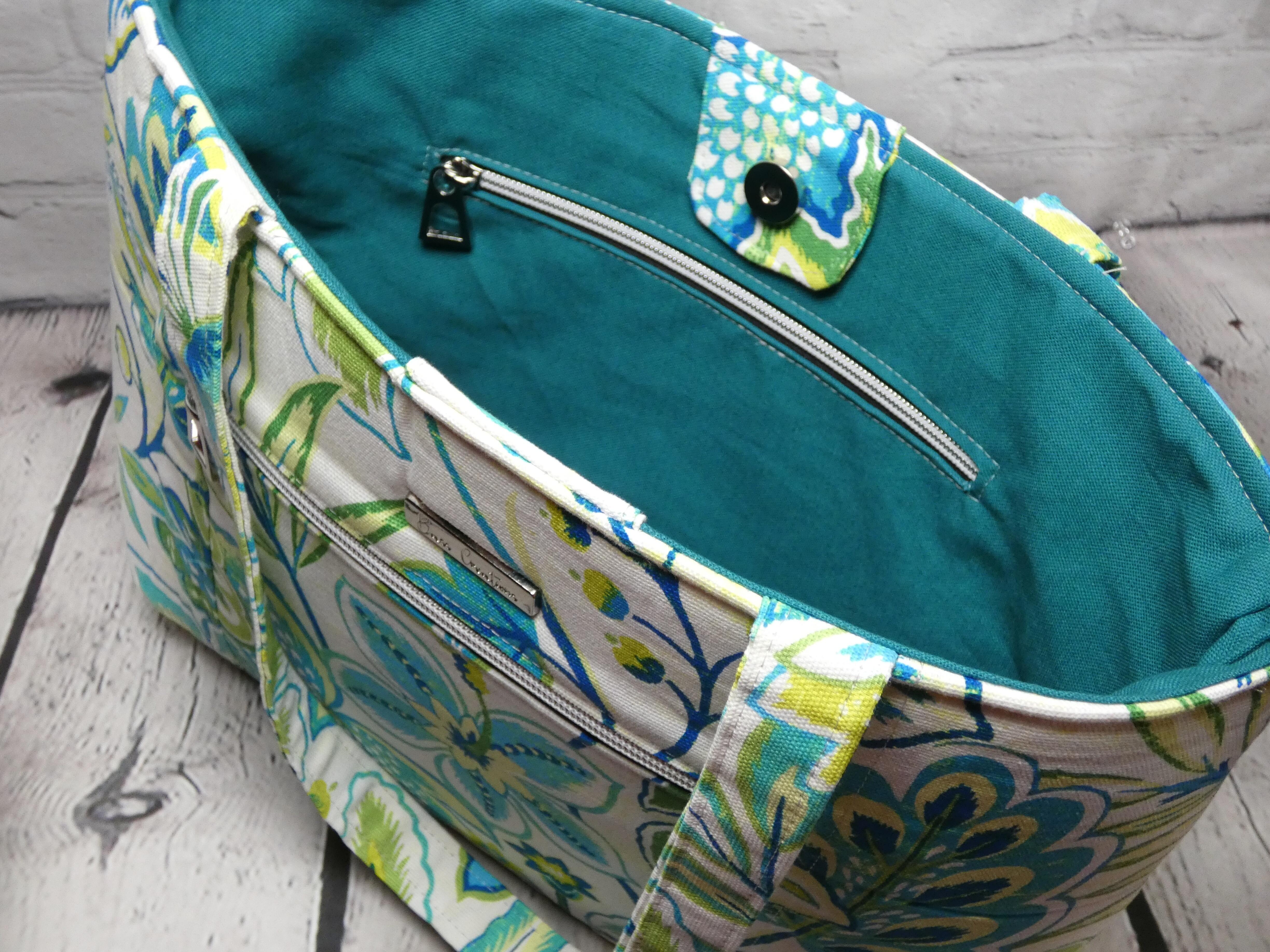 Top view of greens and blues floral pattern market tote with teal lining and an interior zipper pocket, front silver hardware zipper and silver metal Bass Creations logo.
