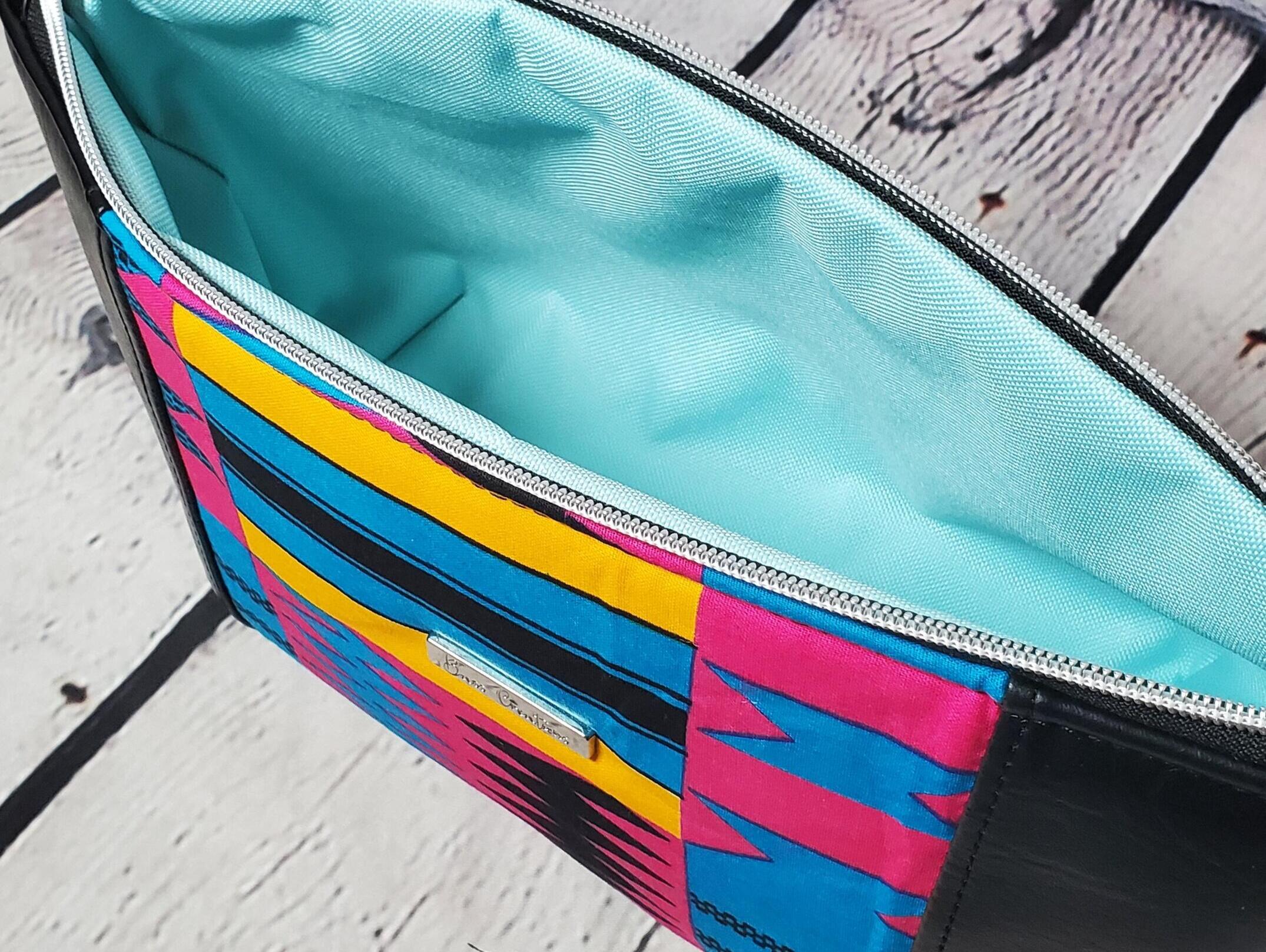 Pink blue and orange geometric print makeup bag with black faux leather accents, silver top zipper and silver Bass Creations logo. Aqua waterproof canvas lining.