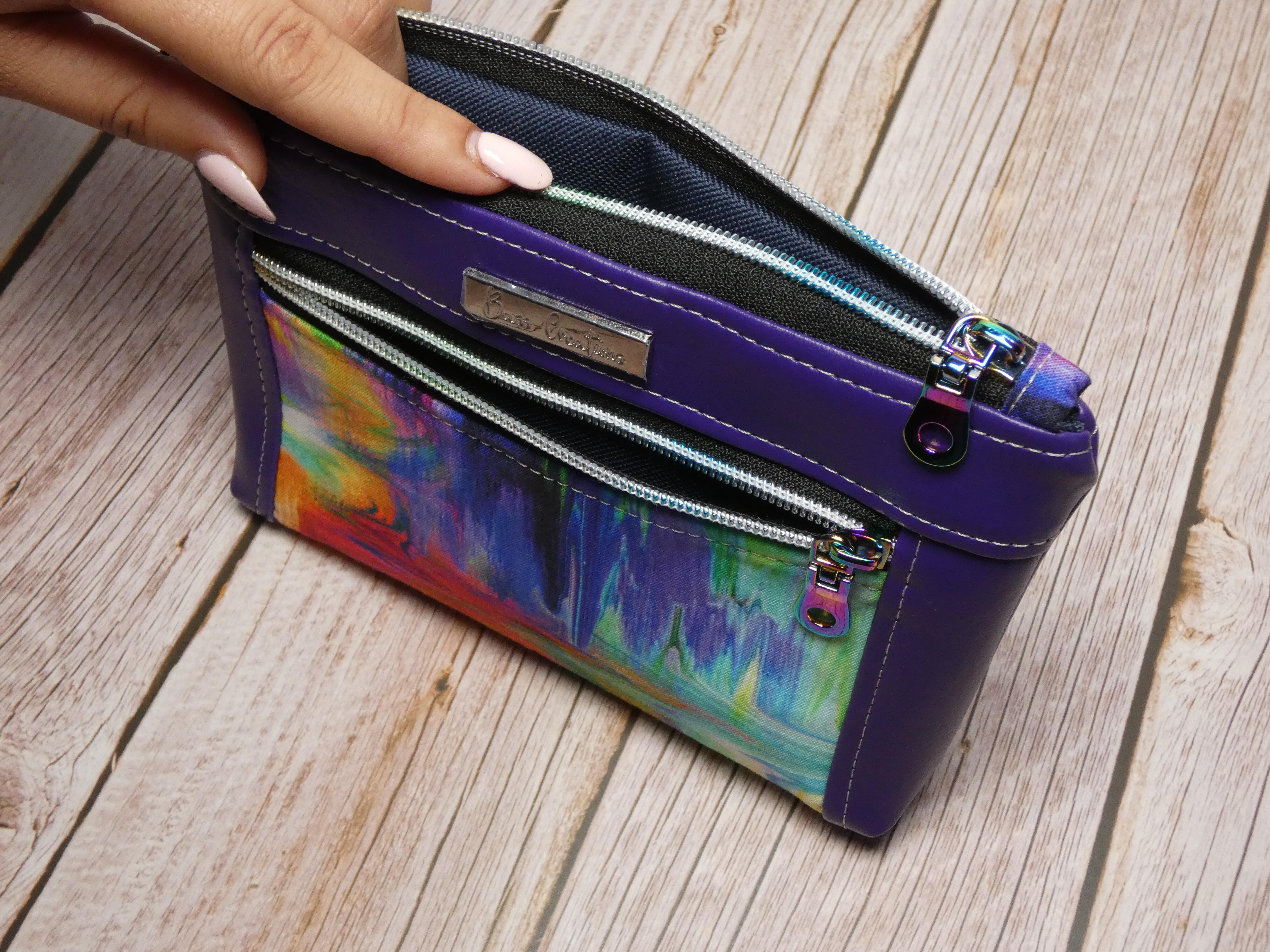 Top view of of a medium makeup pouch with a colorful blurred print fabric and purple faux leather accents. Also showing are the silver toned zipper teeth with silver zipper pulls.