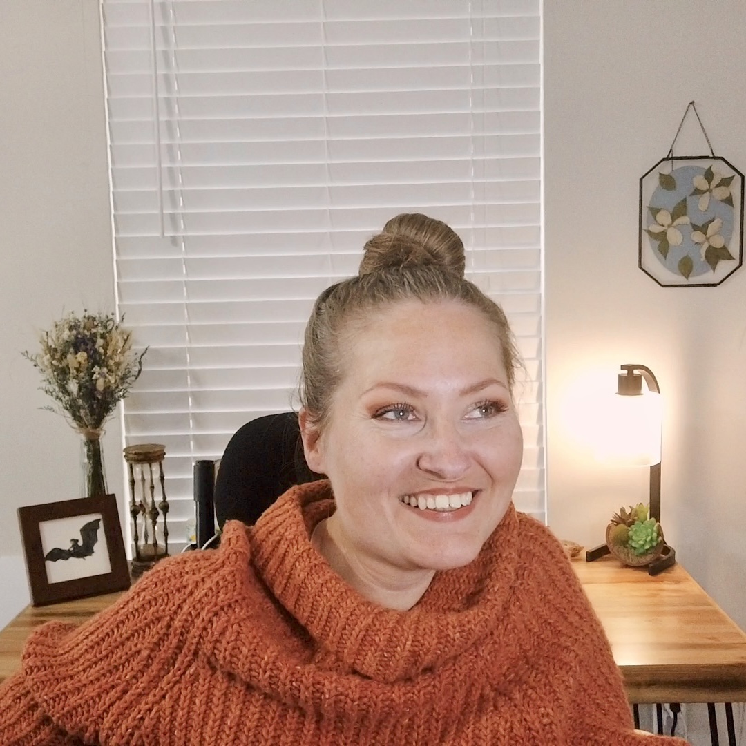 Randi-Lynn looking off to the right smiling with her hair in a top bun wearing a cozy burnt orange sweater. There is a window and desk with decor in the background.