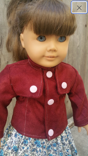 An American Girl doll with brown hair in a ponytail and blue eyes wears a burgundy jacket in front of a wooden fence.