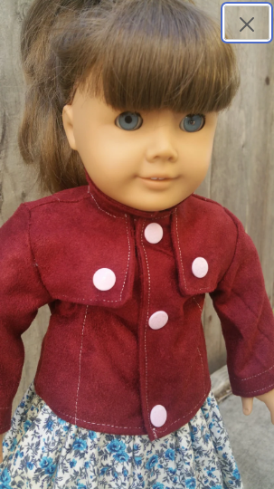 An American Girl doll with brown hair in a ponytail and blue eyes wears a burgundy jacket in front of a wooden fence. An American Girl doll with brown hair in a ponytail and blue eyes wears a burgundy jacket in front of a wooden fence.