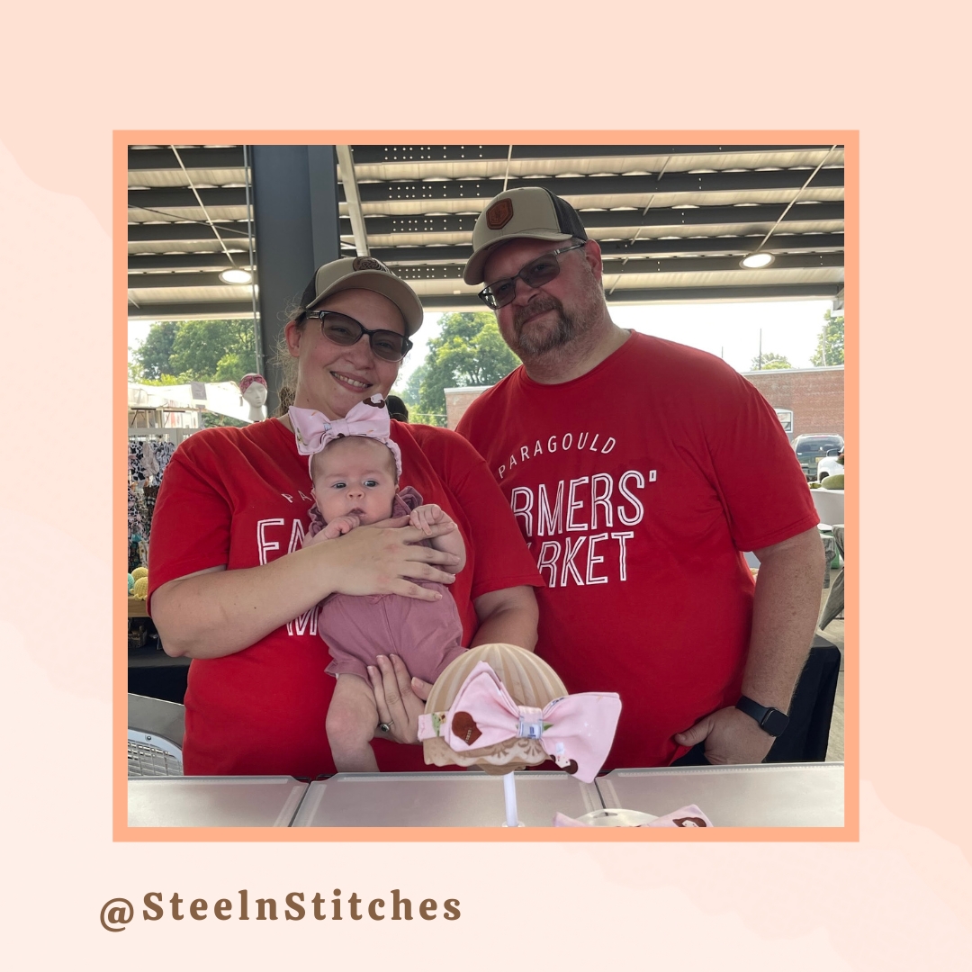 A loving couple wearing matching 'Paragould Farmers' Market' shirts pose happily while holding their adorable baby. The baby is dressed in a cute pink outfit with a big bow headband. A bow sits on the display table in front of them. The backdrop of the market stalls adds to the charming, community feel.
