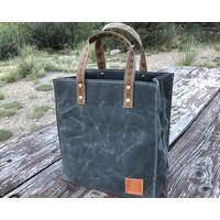Small, stout and sturdy gray waxed canvas tote bag on a wood picnic table in a park. The bag is made in a tiny workshop on wheels. Small, stout and sturdy gray waxed canvas tote bag on a wood picnic table in a park. The bag is made in a tiny workshop on wheels.