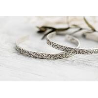 A pair of sterling silver cuff bracelets with an embossed floral pattern, displayed on a rustic white wooden surface. A pair of sterling silver cuff bracelets with an embossed floral pattern, displayed on a rustic white wooden surface.