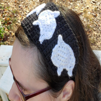 A woman with brown hair wearing a crochet headband with a black band and white ghost appliques. There are brown leaves in the background. A woman with brown hair wearing a crochet headband with a black band and white ghost appliques. There are brown leaves in the background.