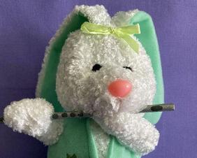 Plush Easter Bunny Flutist made of white chenille and wearing a green vest decorated with carrots, black bead eyes, chiffon yellow hair bow, and pink jelly bean like nose. SO CUTE. Playing a silver wood flute. An ideal basket stuffer for the flutist in your life. She is a sitter for easiness of placement.