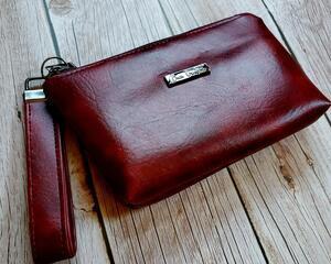 side view of a burgundy marbled faux leather makeup bag with gunmetal hardware and a gunmetal Bass Creations logo. It also includes a wristlet strap for easy carrying.
