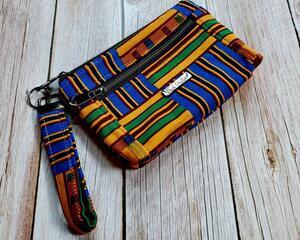Side view of a golden yellow, green and blue Kente printed makeup pouch with 2 gunmetal toned zipper pockets and a gunmetal toned Bass Creations logo.