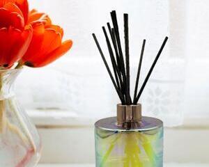 Iridescent oil diffuser bottle with rose gold cap and black reeds. On table next to vase with flowers.