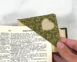 Fabric corner bookmark made from tan and olive green fabrics with a heart applique on the front. It is being slid over the corner of a page to demonstrate how to use this bookmark.