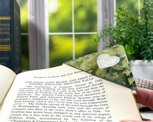 A green and cream colored fabric corner bookmark with a cream applique heart on the front is shown on a page of a book with a window and plant in the background of the photo.