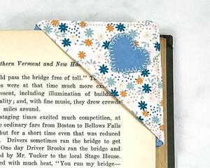 A fun blue, peach color and white fabric corner bookmark with a blue heart applique.  The corner bookmark is being shown slid over a page of a book to show it in use.
