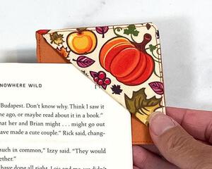 A fun fall fabric design was used to make a corner bookmark with leaves, pumpkins, etc.  The bottom of the bookmark is orange and the picture depicts it being slid over a page in a book to demonstrate how to use this handmade bookmark corner.