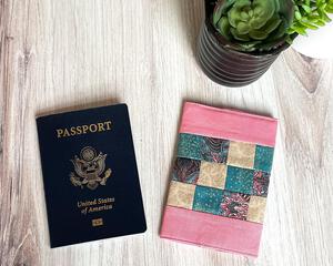 A pink green and tan fabric patchwork passport cover is lying on a table with a passport displayed beside it and some succulent plants at the right of the photo.  These covers perfectly wrap around all passports to add a splash of style and uniqueness to an otherwise boring travel document.