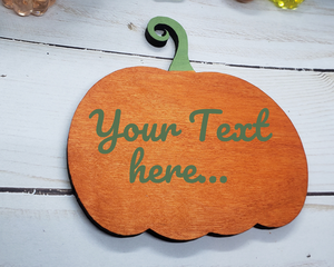 A wooden pumpkin magnet painted in a subdued shade of orange with a sage green curly stem. The same sage green is printed across the pumpkin and says "Your text here.."