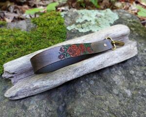 Classic red roses with deep green foliage adorn this handmade leather wrist strap, which was cut, etched, dyed and painted right here in our shop. Made from incredibly strong and durable vegetable tanned full grain leather, the wrist strap is hand dyed in Antique Black with a slightly distressed or vintage look.