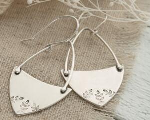 Handcrafted sterling silver fern-stamped hoop earrings on burlap with white flowers, made in Soldotna, Alaska. 

