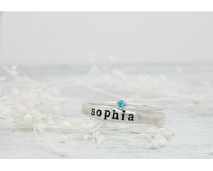 Sterling silver name ring with "Sophia" and blue birthstone ring on white floral background.
