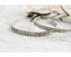 A pair of sterling silver cuff bracelets with an embossed floral pattern, displayed on a rustic white wooden surface.