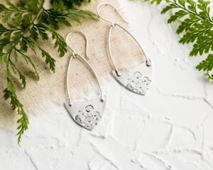 Handcrafted sterling silver Art Nouveau floral hoop earrings on a textured background with greenery.
