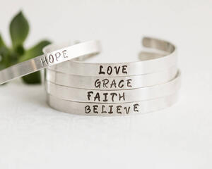 Five sterling silver bracelets with words HOPE, LOVE, GRACE, FAITH, BELIEVE, on a white surface with a green succulent in the background.

