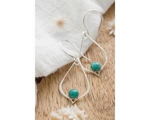 "Pair of silver drop earrings with turquoise gemstones, resting on a white fabric with dried flowers on a wooden surface."