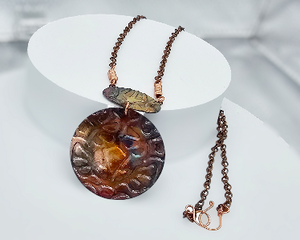 Round copper pendant colored in deep shades of red, blue, yellow created by flame coloring hanging under oval copper piece on copper chain