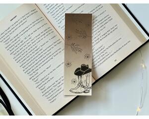 this western bookmark has ombre brown background with cowboy boots with a hat on top of the boots. there are flowers all over the book mark