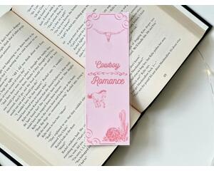 this cowboy romance bookmark has a pink background with a little bit of a darker pink boarder on top and bottom left. there is a cactus in the bottom right. In the middle of the bookmark there are words Cowboy Romance with a cute design between