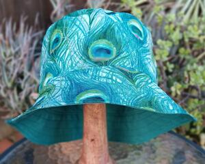 Front view of bucket hat on stand. All over print features drawn peacock feathers on light blue background. Part of the reverse side of hat is visible to show the coordinating blue-green.