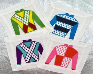 4 white linen cocktail or beverage napkins/coasters  with different multi-colored jockey silks on each laying on a silver background. 