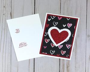 A valentine card making kit of 12 with black scrapbook paper with hearts, white and red heart embellishments, and white cards and envelopes.
