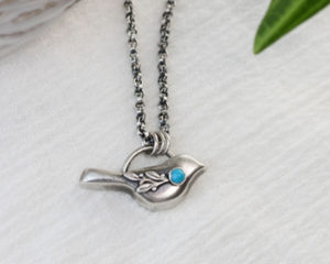 Sterling silver bird necklace with turquoise, on a white surface with green leaves and a decorative bowl.

