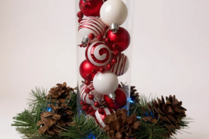 Handmade Christmas centerpiece with red and white swirl ornaments in glass vase, pine greenery, pinecones and blue light on wood slice base
