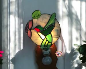 Hummingbird stained glass night light- made of green white and red glass, background is of light blue glass with a pink flower and lampworked green leaf. Show with light on.