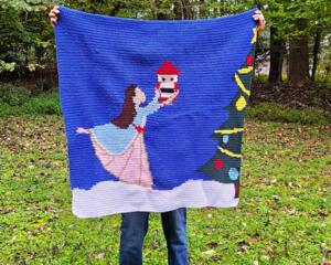 A woman holding a crochet baby blanket with a picture of a ballerina dancing with a Nutcracker doll in the snow on it. 