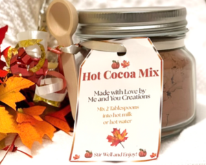 Handmade hot cocoa mix jar with wooden spoon and red-gold ribbon, displayed on a white knit blanket with autumn leaves.