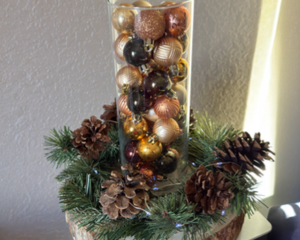 Rustic holiday centerpiece with pinecones, greenery, and a glass vase filled with gold and bronze ornaments on a wood-slice base.