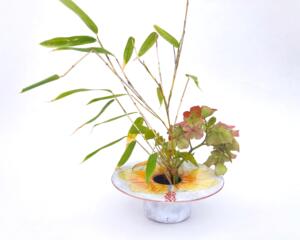 photo of kiln-fired copper enamel ikebana-style vase with hand painted hibiscus on top with is 4 inches in diameter. Enamel colors of hibiscus are pale yellow to orange, with pale green leaves. Vase stands 1-1/4 inch tall. There is a pin-frog inside to hold flowers upright.
