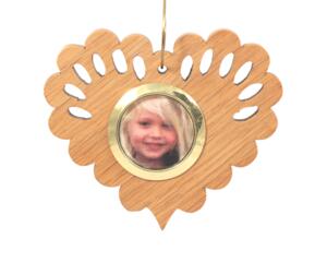 Wooden heart-shaped ornament with scalloped edges and an oak grain finish, featuring a removable round gold frame that holds a photo of a smiling blonde child.
