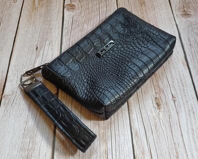 side view of a black alligator faux leather makeup bag with gunmetal hardware and a gunmetal Bass Creations logo. It also includes a wristlet strap for easy carrying.